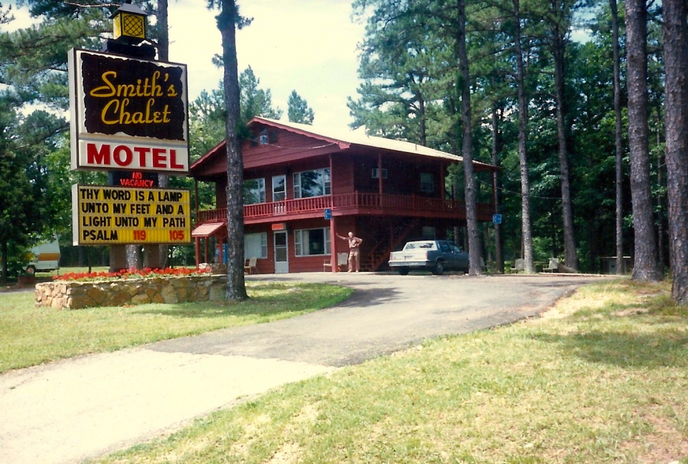 Buel Smith leaning against post of motel behind large Smith's Chalet sign.