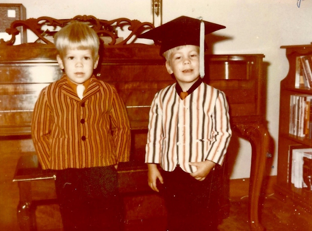 Two pre-school boys dressed in their Sunday best standing in front of an ornate piano with one wearing his father's graduation cap.