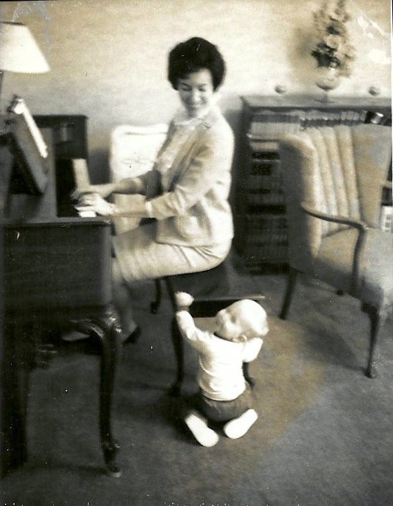 A young mother playing piano as her 10-month old son plays near her.