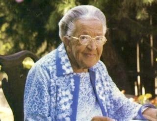 The older lady, Corrie ten Boom, in a blue and white dress sitting in a chair outside.