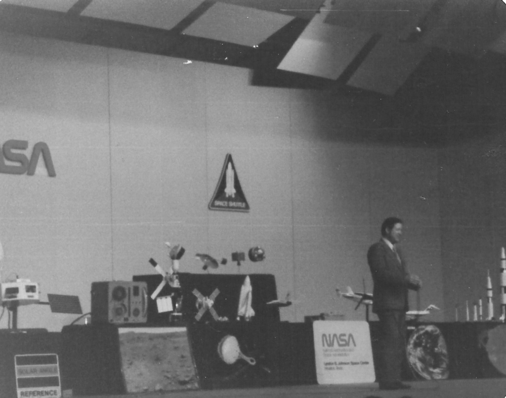 A man in a suit on the stage in the NASA auditorium with lots of model space ships on the table behind him.