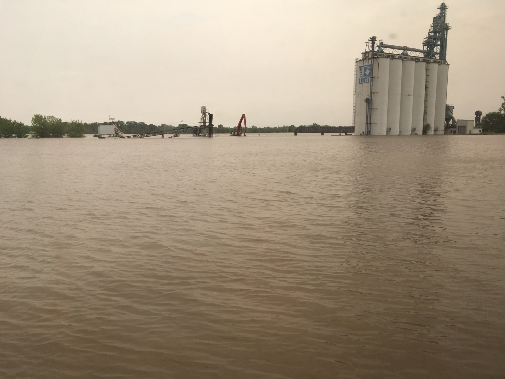 View seen from inside the train of large flooded area beside the train tracks showing farm equipment and grain silo standing in water.