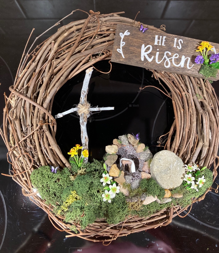 View of Easter wreath showing a wooden cross to the left with an empty tomb below it on the right. Flowers and butterflies abound across the bottom.