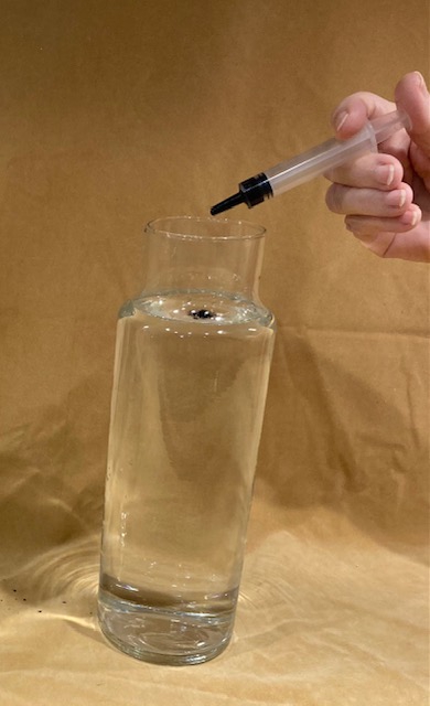 A hand dropping one drop of black ink into a glass of water.