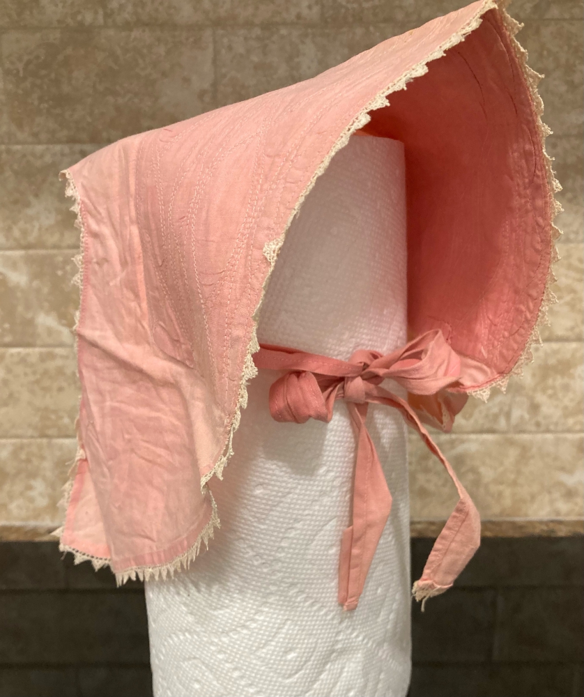 A small pink sun bonnet trimmed in white lace tied with a bow on a roll of paper towels head.