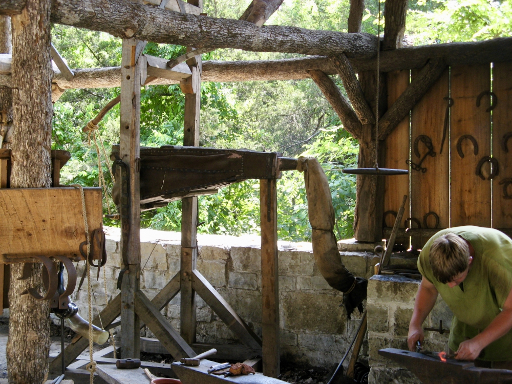 Blacksmith bent over anvil hammering on glowing hot piece of metal with bellows for adding air to the forge are in the background.