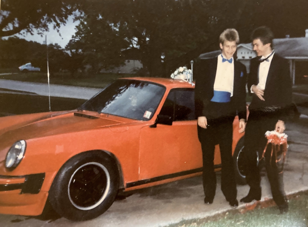 Two teenage boys in tuxedos standing beside red Porsche 911 car.