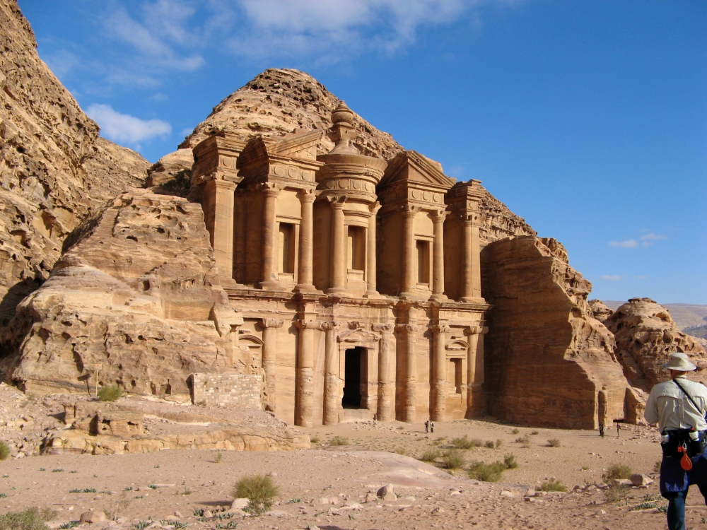 Front of Petra's Monastery that is carved out of sandstone and 160 feet tall showing two people in front barely tall enough to come up to the base of the 26 foot tall door opening. 