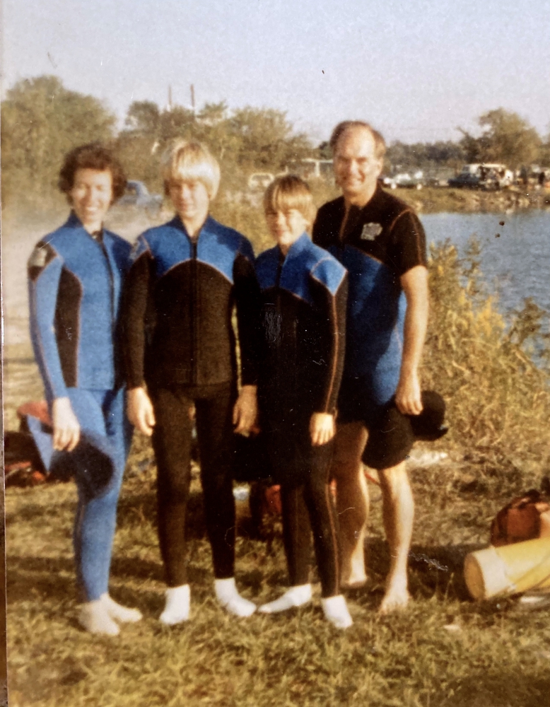 Husband, wife and 2 sons in wet suits beside water.