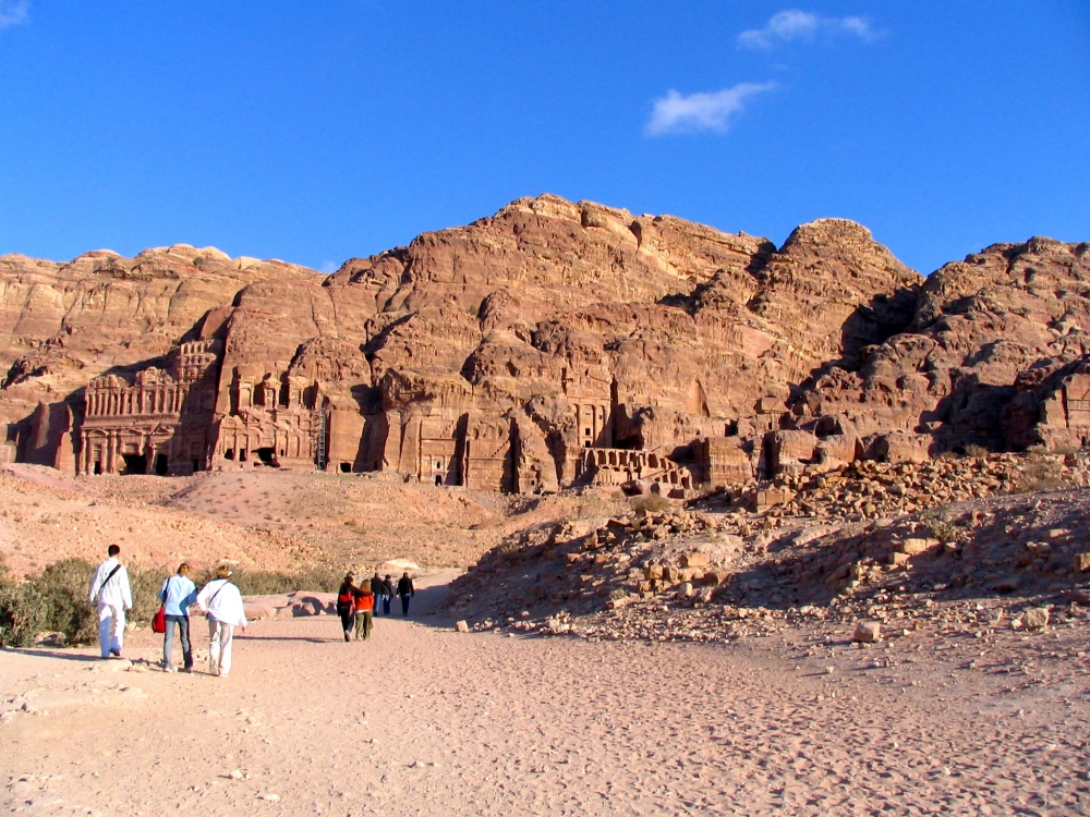 Several people  walking across flat area inside Petra toward mountains in background with carved tombs and buildings seen carved into mountains.